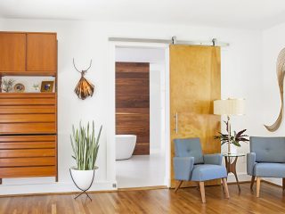 Image shows a white room with a sliding barn door. There's a floor lamp between two chairs and modern wall art.