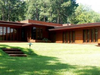Image shows the front exterior of a ranch house with floor to ceiling windows surrounded by trees and grass.