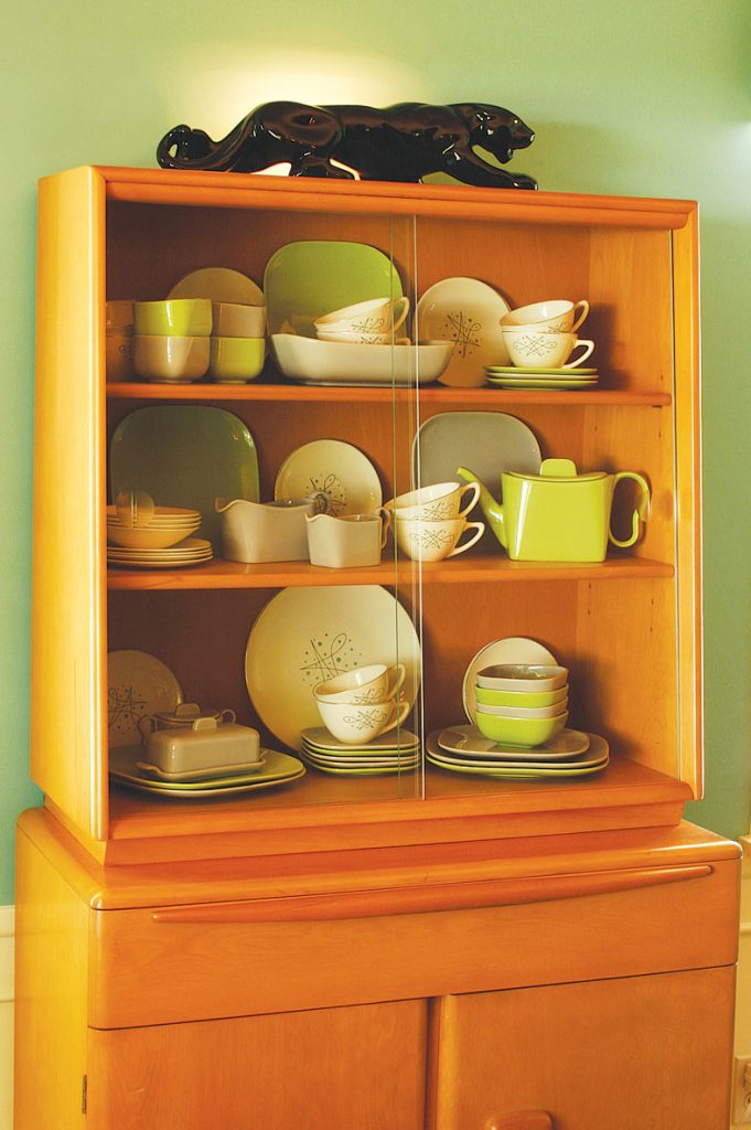 A blond china hutch showcases green and cream dishware. A black panther light sits on top