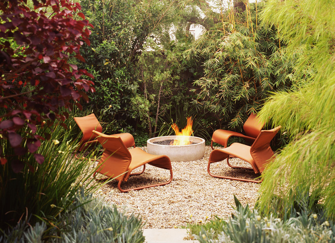 A backyard fire area with a round fire pit and two orange chairs, surrounded by plants and trees