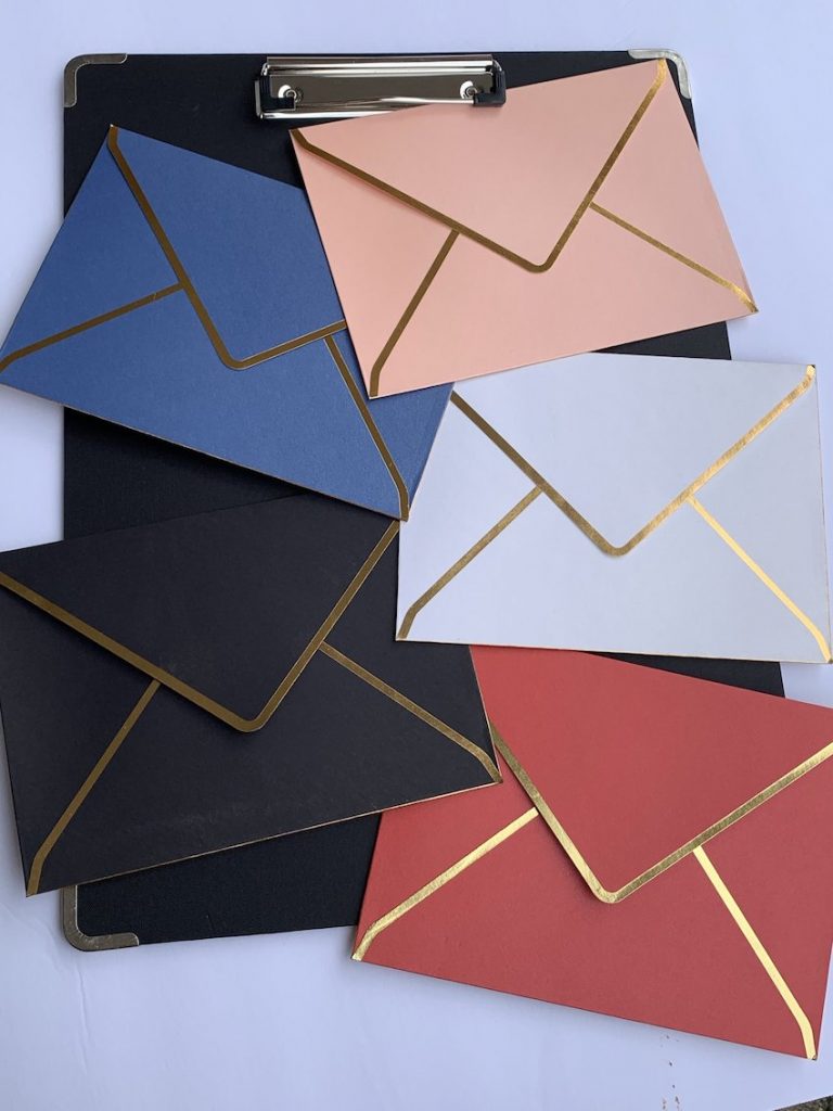 5 envelopes with gold trim in different colors are arranged on a black clipboard