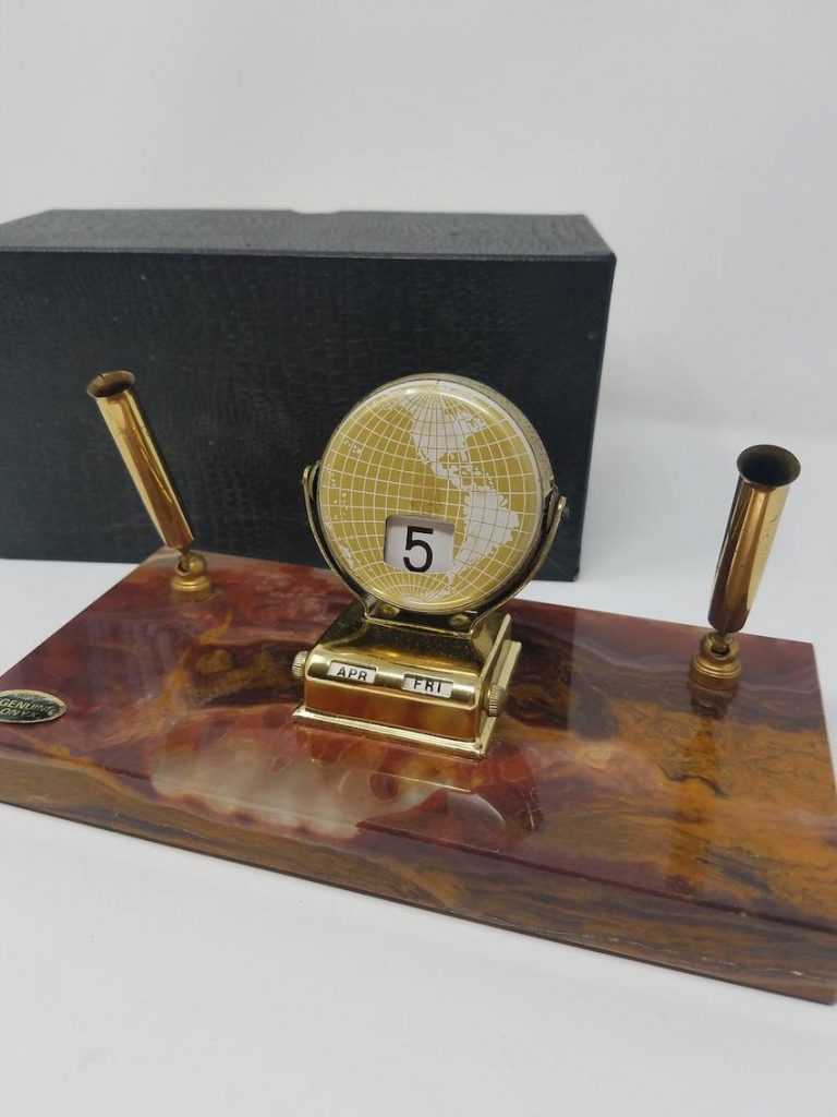 A desk calendar on a marble base with two gold pen holders and a flat gold globe with a flip calendar.