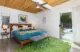 A bedroom with a wooden mid century modern style bed frame, blue bedding and a colorful throw blanket folded at the end of the bed. there is a sliding glass door which leads to the backyard which has a pool. 