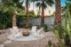 A round fire pit with 4 modern chairs arranged in a circle around it. The area sits on a round cement pad which is surrounded by gravel and bordered by agave plants and palm trees