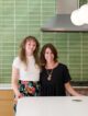 two women in a green tiled kitchen standing behind a white countertop.