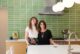 two women in a green tiled kitchen standing behind a white countertop.