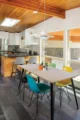A small dining room of a mid century modern home. there are wood planked ceilings and mid century modern hanging lamps above colorful molded plastic chairs. In the background is a simple mid century modern style kitchen.