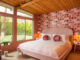 bedroom with modern pink geometric wallpaper an upholstered bed with white sheets and two decorative round throw pillows. The ceiling of the room is a wood plank.