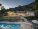 Twilight view of a mid-century modern home with warm interior lighting glowing through floor-to-ceiling glass walls, rooftop solar panels, a freeform pool in the foreground, terraced gardens with stone staircase, raised garden beds, and the San Gabriel Mountains as a backdrop