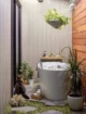 "Spa-like indoor-outdoor bathroom nook featuring a deep round white Japanese soaking tub with a floor-mounted brushed nickel faucet, a floating wood bath shelf with candles and bath products, a hanging pothos plant in a woven basket, horizontal wood plank accent wall, vertical shiplap painted walls, irregular flagstone flooring with moss ground cover, lush potted tropical plants, pillar candles on a wood tray, a coral sculpture, and a tabby cat sitting by the glass door.