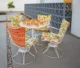 Vintage white wire swivel patio dining set with bold orange, green, and white mod-print cushions, set with orange tiki glasses on a round mesh table, positioned in front of a decorative gray breeze block screen wall on a concrete patio with gravel planting beds