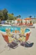 Vintage mid-century modern poolside patio with white wire swivel chairs cushioned in a bold orange, green, and white geometric print, surrounding a round mesh table set with orange tiki glasses, beside a kidney-shaped pool with orange chaise lounges and matching umbrellas, backed by tall saguaro cacti and palm trees under a deep blue desert sky