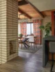 "Mid-century modern dining room viewed past a floor-to-ceiling brick fireplace column with stacked firewood, featuring a walnut oval dining table with an angular black metal base, walnut dining chairs with dark upholstered seats, bold orange and gray geometric print curtains, a woven grasscloth accent wall, a gray jute area rug, a wrought iron drum pendant light, a vaulted wood-planked ceiling with exposed dark beams, a fiddle-leaf fig plant, and an ocean glimpse through the window"