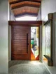Mid-century modern entryway featuring a solid horizontal-plank walnut pivot door with a tall black bar handle, an adjacent stained glass sidelight panel with geometric shapes in amber, green, red, and clear glass, a vaulted wood-planked ceiling with skylight above, a brass wall sconce, a live-edge wood sculpture, and a round patterned area rug on hardwood floors