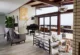 A living room in a mid-century modern home renovation with a vaulted wood-planked ceiling, exposed dark wood beams, a black baby grand piano, a built-in cushioned hearth seating area with white chevron pillows beside a brick fireplace column, floor-to-ceiling sliding glass doors opening to a wood deck with ocean views, a sculptural glass pendant light, a gray accent chair with throw blanket, an arc floor lamp, and a gold abstract circular artwork on the wall"