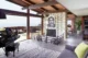 Mid-century modern living room with a vaulted wood-planked ceiling, exposed dark wood beams, a black baby grand piano, a two-sided brick fireplace with black iron surround, a large-scale black and white portrait artwork, floor-to-ceiling sliding glass doors opening to a wood deck with ocean views, a gray shag rug, gray sofa with yellow accent pillow, and a kitchen visible in the background