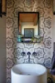 "Powder room featuring a floor-to-ceiling Moroccan-style geometric patterned encaustic tile wall in cream, gray, and navy, a round white pedestal vessel sink, wall-mounted navy industrial-style wheel handle faucet, a black-framed recessed mirror with a small metal shelf holding hand soap and a succulent, and brass wall sconce accents flanking the mirror"
