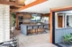 A mid-century modern home renovation with a did-century modern open kitchen with walnut flat-front cabinetry, gray subway tile backsplash, stainless steel double wall ovens, a gas cooktop, stainless French door refrigerator, a large concrete-topped island with four gray upholstered metal bar stools, a sculptural blue pendant light, wood-look tile floors, exposed orange-painted wood beams, a brick column accent wall, a fiddle-leaf fig plant, and folding glass doors opening to a planted atrium"