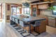 A kitchen in a mid-century modern home renovation featuring a large concrete-topped island with walnut base and four gray upholstered metal cantilever bar stools, walnut flat-front cabinetry, gray subway tile backsplash, stainless steel double wall ovens, a gas cooktop, stainless dishwasher, a sculptural blue pendant light above the island, a wrought iron drum pendant light, wood-look tile floors, orange-painted exposed wood beams, a brick fireplace column, trailing pothos plants, and windows with views to a wood deck beyond"