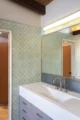 "Mid-century modern bathroom featuring a floor-to-ceiling sage green geometric patterned encaustic tile accent wall, a white rectangular integrated sink with brushed nickel widespread faucet, a periwinkle blue vanity with brushed nickel hardware, a frameless wall mirror with a linear vanity light, and an exposed dark wood ceiling beam above"