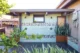 Outdoor shower area on a mid-century modern home's exterior, featuring a mint green brick wall with a decorative mosaic tile accent band, two wall-mounted shower fixtures, a small black-framed window, a warm wood door, built-in wood planters with snake plants and tropical foliage, dark slate tile flooring, and a vaulted wood-planked overhang with exposed beams.