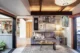 Mid-century modern living room in a mid-century modern home renovation with a wood-planked coffered ceiling, exposed dark steel beams, a gray leather sofa with neutral pillows, a live-edge epoxy resin coffee table, an eclectic gallery wall of nature-themed framed artwork, a linen drum floor lamp, a vintage-style area rug, and folding glass doors on both sides opening to an atrium and outdoor space, with a kitchen visible in the background