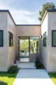 Minimalist modern home entry courtyard with warm taupe plaster walls, flat roofline with dark fascia, black-framed windows, a concrete stepping stone path flanked by low plantings, a black dome wall sconce, and a black-framed sliding glass door opening to a view of an outdoor dining area beyond