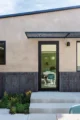 Modern home exterior detail with warm taupe plaster upper walls, a dark charcoal running-bond brick wainscot, a full-glass black-framed entry door with perforated metal canopy overhead, a brass and black wall sconce, concrete steps, and a low-water succulent garden planting at the base