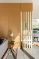 Minimalist Malibu mid century modern interior with a floor-to-ceiling warm oak panel wall and vertical wood slat room divider casting geometric shadows on a polished concrete floor, beside a light wood tripod side table with a small plant and white table lamp, with a gray sofa, vintage kilim rug, and a glimpse of a garden view through a picture window beyond