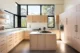 Sun-filled modern kitchen with flat-front light oak cabinetry, a central island with white quartz countertop, gas cooktop, floating wood shelves, blush pink tall pantry wall, and a wraparound band of black-framed windows flooding the space with natural light and treetop views, on polished concrete floors