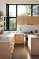 Bright modern kitchen with flat-front light oak cabinetry, white quartz countertops, stainless steel professional range, floating wood shelves, and dramatic black-framed floor-to-ceiling windows framing a treetop view, on polished concrete floors