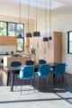 Open-plan dining area with a dark wood dining table surrounded by teal velvet Beetle chairs, lit by a cluster of matte black pendant lights, opening to a modern kitchen with light oak cabinetry, blush pink tall pantry cabinets, and black-framed windows framing a treetop view, on polished concrete floors
