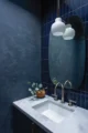 Moody powder room with deep navy blue venetian plaster walls, vertical navy ceramic tile backsplash, a marble undermount sink with brushed nickel faucet, rounded frameless mirror, a globe pendant light with brass stem, and a small eucalyptus arrangement in a wood bowl