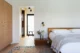 Serene modern bedroom with a wood platform bed dressed in white linen, a dark walnut nightstand with a small floral arrangement, brass wall sconce, built-in light oak dresser, vintage Persian rug, and a view through an open doorway into a walk-in closet with black-framed window