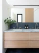Modern double vanity bathroom with a floating light oak cabinet, honed marble countertop with dual undermount sinks and brushed nickel faucets, slate blue vertical tile backsplash, full-width frameless mirror with black bar light, dark tile floor, and a glass vase of eucalyptus branches