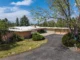 Aerial view of a mid-century modern ranch home on a corner lot with a long asphalt driveway, pink brick and stacked stone facade, a low-pitched roof, mature trees including blue spruce and deciduous trees, manicured shrubs, and a large lawn