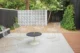 Backyard patio with a patterned breezeblock wall, round outdoor table, white chairs, concrete paving and mature landscaping.