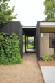 Side entrance to the Mid-Century Modern home designed by Victor Hosford with a black exterior, concrete walkway, gravel landscaping, mature greenery and large windows.