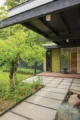 Black-painted 1958 Mid-Century Modern home with a covered entry, gravel courtyard, stepping-stone path, mature trees and a green front door.