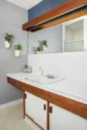 Updated bathroom vanity with white countertop and sink, wood cabinetry, blue-gray wall, white tile and floating wood shelves with plants.