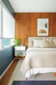 Primary bedroom with a white bed, mahogany-paneled accent wall, blue-gray walls, large windows and a small bedside table.