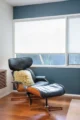 Mid-Century Modern lounge chair and ottoman beside a large window, with blue-gray walls, wood flooring and a small side table.