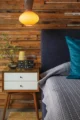 Bedroom detail with a dark upholstered headboard, textured gray bedding, blue pillow, wood-paneled wall, yellow pendant light and vintage bedside clock.