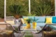 Outdoor seating area with a turquoise sofa, woven chairs, yellow cushions and potted plants beside a lush hedge.