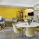Mid-Century Modern living and dining area with a bright yellow wall, round dining table, olive chairs, white sofa and large glass doors.