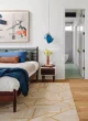 Mid-Century Modern bedroom with a wooden bed, blue and patterned pillows, blue pendant light, abstract artwork, wood flooring and a cream rug with a geometric pattern.