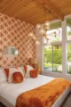 Mid-Century Modern bedroom decor ideas with a white bed, orange velvet pillows and throw, geometric patterned wallpaper, wood-plank ceiling, globe pendant light and large windows overlooking a garden.
